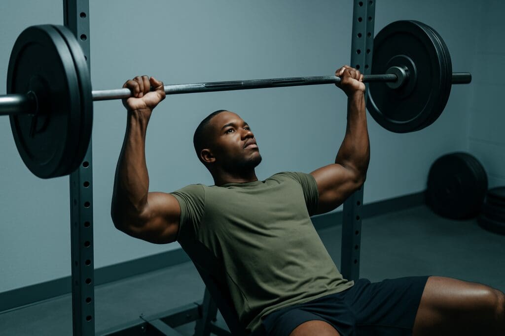 A strong, focused man performs a barbell bench press in a minimalist gym, his defined upper body and olive-green shirt accentuated by soft, even lighting. The sleek setting and intense posture highlight the performance health benefits of strength training for muscular power and control.